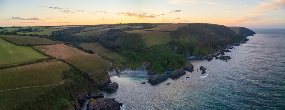 Aerial view of Lansallos Beach with rocky cliffs and green coastal fields illuminated by golden hour light in Looe, Cornwall, United Kingdom.