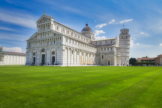 Pisa, Italy - 03 May 2022: View of the Leaning Tower of Pisa and the Cathedral of Santa Maria Assunta set against a vibrant blue sky with a lush green lawn in the foreground.