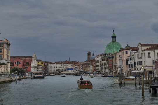Venice, Veneto, Italy - 06 May 2022: View of the Grand Canal in Venice with a wooden water taxi moving past historic buildings and the prominent green dome of San Simeone Piccolo under a cloudy sky.