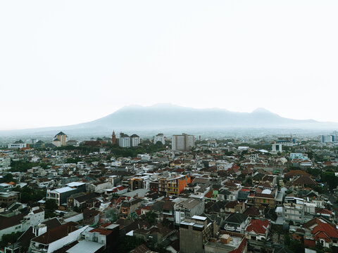 Aerial view of dense residential rooftops and urban buildings with a large mountain looming in the misty background in Malang, Jawa Timur, Indonesia.