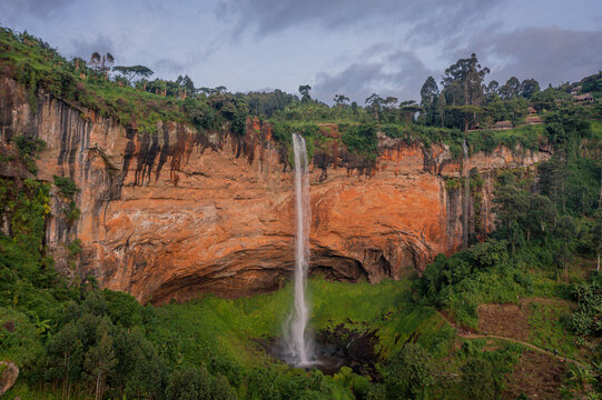 Aerial view of Sipi Falls cascading down a dramatic orange-hued cliff face surrounded by lush green vegetation and trees in Kapchorwa, Eastern Region, Uganda.