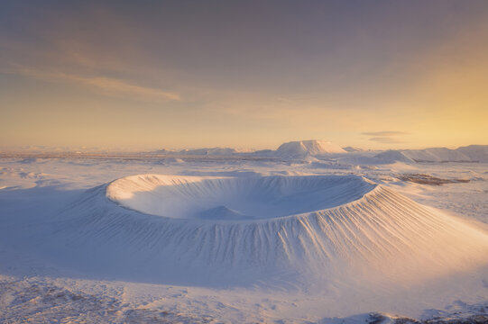 Aerial view of a snow-covered volcanic crater in a vast icy landscape illuminated by the warm light of the golden hour in Iceland.