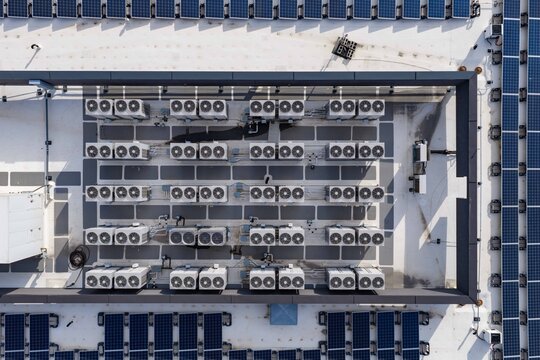 Aerial view of rows of industrial air conditioning units and ventilator fans on a flat office rooftop surrounded by solar panels in Lehi, Utah, United States.