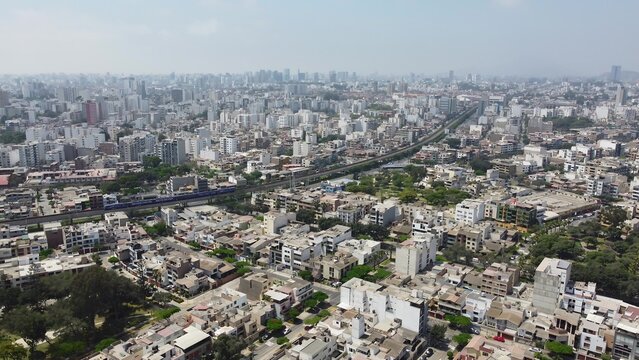 Aerial view of the dense urban landscape featuring residential buildings, a railway line, and green parks under a clear sky in Santiago de Surco, Lima Province, Peru.
