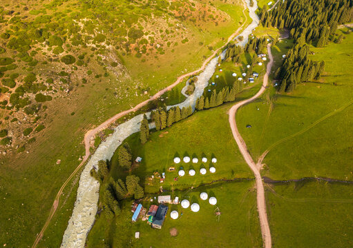 Aerial view of Kok Jaiyk valley featuring a winding river, lush green meadows, pine forests, and a traditional yurt camp in Dzhetyoguz, Issyk-Kul Region, Kyrgyzstan.