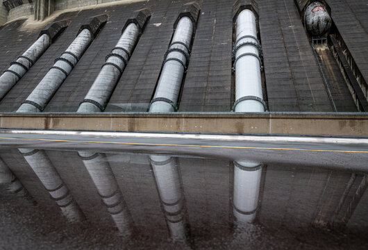 View of the Revelstoke Dam with its massive penstocks and concrete structure reflected in a puddle on the access road in Revelstoke, British Columbia, Canada.