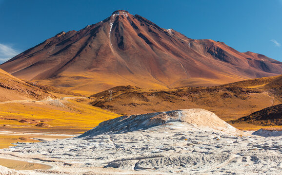 View of Piedras Rojas with its distinctive white salt formations and the towering reddish volcanic mountain under a clear blue sky in San Pedro de Atacama, Antofagasta, Chile.