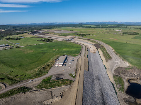 Aerial view of the Elbow River flood mitigation project featuring large rock embankments, dirt roads, and surrounding green fields under a clear blue sky in Calgary, Alberta, Canada.
