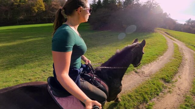 SELFIE, LENS FLARE: Young woman rides her dark brown horse along scenic dirt path beside an open meadow. Warm autumn sunlight shines through forest trees as they trot through tranquil countryside.