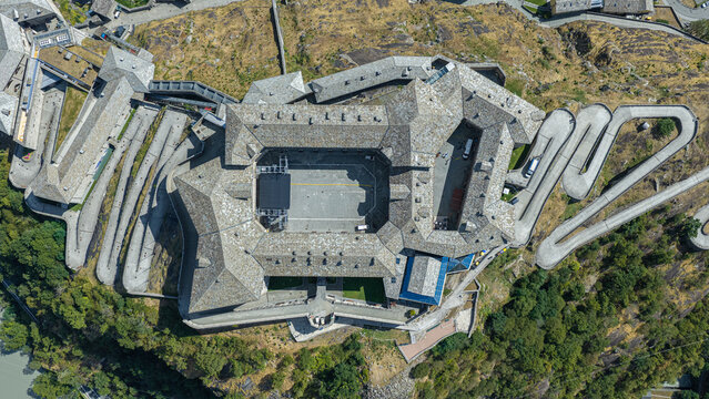Aerial view of Forte di Bard with its historic stone rooftops, central courtyard, and winding access roads surrounded by rocky terrain and lush greenery in Bard, Valle d'Aosta, Italy.