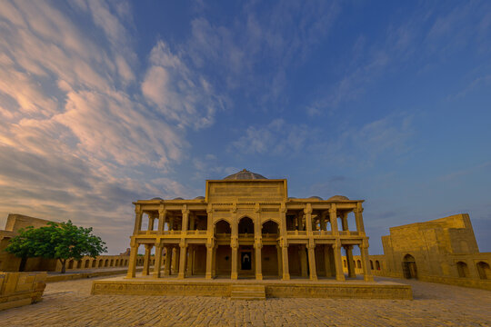 View of the historic Isa Khan Tarkhan Tomb with intricate stone carvings and arched colonnades under a dramatic golden hour sky in Thatta, Sindh, Pakistan.