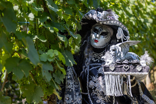 Riquewihr, France - 04 July 2026: View of a person in an elaborate Venetian masquerade costume with a silver mask and ornate clothing, holding a winged lion figurine in a vineyard.