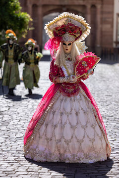 Riquewihr, France - 04 July 2026: View of a person dressed in an elaborate red and white Venetian carnival costume with a mask and fan, walking on a cobblestone street in Riquewihr.