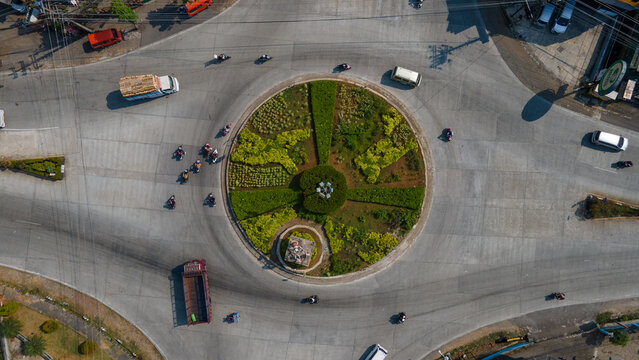 Aerial view of a roundabout with landscaped greenery and a central monument, featuring traffic including cars, trucks, and motorcycles in Banyumas Regency, Central Java, Indonesia.
