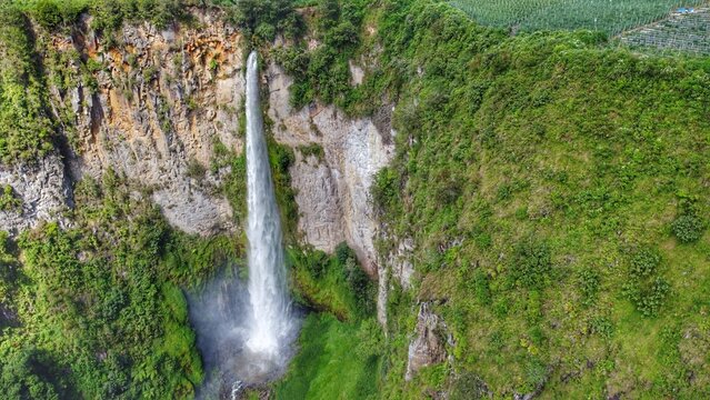 Aerial view of Sipiso Piso Waterfall cascading down a lush green cliff face with rocky textures and dense vegetation in Merek, North Sumatra, Indonesia.
