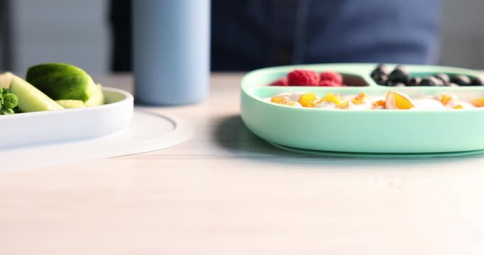 Baby tableware. Woman putting section plate with food onto light wooden table, closeup