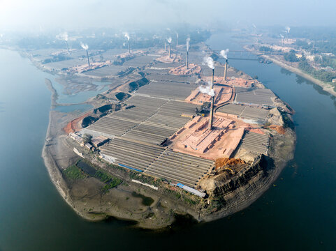 Aerial view of a brick manufacturing site with tall chimneys emitting smoke, rows of drying bricks, and industrial structures on a riverbank in Tangail, Dhaka Division, Bangladesh.