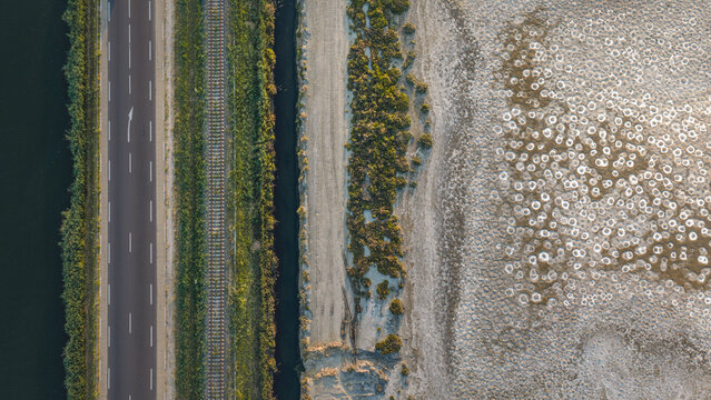 Aerial view of parallel transport infrastructure including a road and railway tracks beside a salt marsh with abstract white mineral patterns on the ground, France.