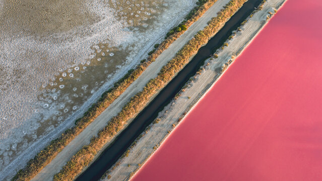 Aerial view of a vivid pink salt pond divided by a dark water channel and narrow embankments from textured saline ground, forming a striking abstract geometric landscape in France.
