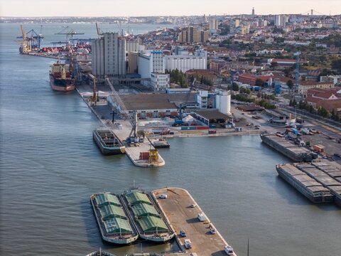 Aerial view of the Braco de Prata industrial port area with silos, cargo barges on the Tagus River, and the urban skyline of Lisboa, Lisboa, Portugal.
