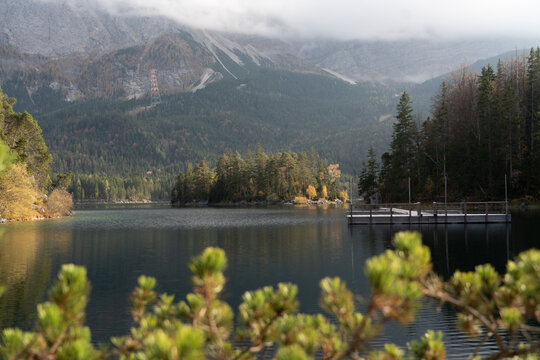 View of the calm Eibsee lake with a wooden pier, an island covered in pine trees, and the towering Zugspitze massif under a cloudy sky in Germany.