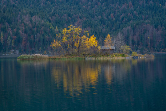 View of a small island with vibrant yellow autumn trees and a rustic wooden cabin situated in a calm lake, backed by a dense forest in Germany.