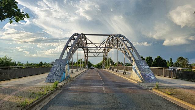 View of the Holzwiesenbrucke steel arch bridge with graffiti on its concrete supports and a dramatic cloudy sky in the background in Hannover, Niedersachsen, Germany.