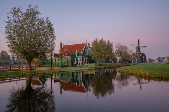 View of a traditional green wooden house along a calm canal with a windmill in the background under a soft sunset sky in the Netherlands.