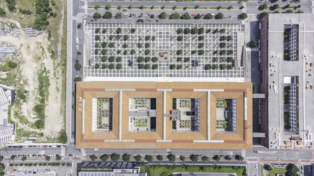 Aerial view of the University of Milano-Bicocca campus, featuring a rectangular building with terracotta roofs and internal courtyards, next to a grid-patterned plaza, in Milan, Lombardy, Italy.