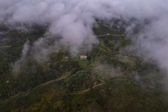 Aerial view of Cape Roca surrounded by lush green hills, rolling terrain, and a solitary house partially obscured by low-hanging clouds in Colares, Lisboa, Portugal.