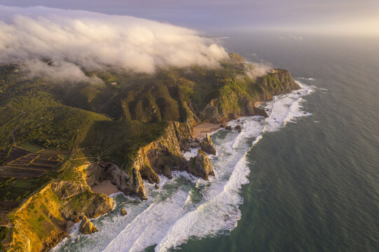 Aerial view of the dramatic cliffs and coastline at Cape Roca, with crashing waves, lush green hills, and low-hanging clouds during golden hour in Colares, Lisboa, Portugal.