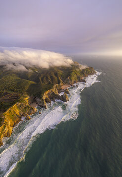 Aerial view of the dramatic cliffs of Cape Roca at sunset, with waves crashing against the rugged coastline and low clouds rolling over the green hills in Colares, Lisboa, Portugal.