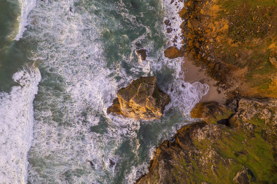 Aerial view of Cape Roca featuring a large rock formation surrounded by crashing waves, rugged cliffs, and golden light in Colares, Lisboa, Portugal.