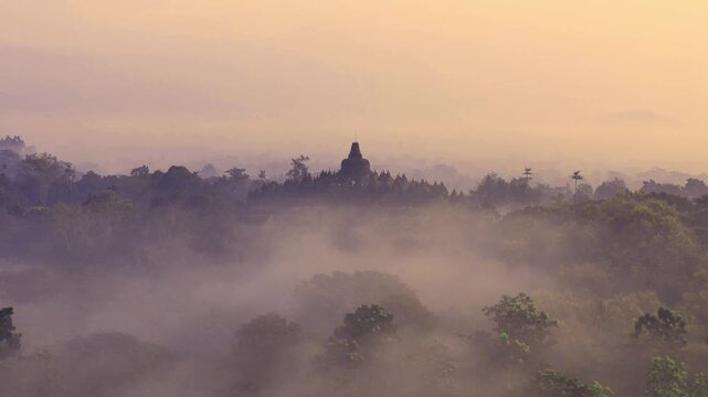 Aerial view of Borobudur Temple surrounded by lush trees and morning mist during sunrise in Kecamatan Borobudur, Jawa Tengah, Indonesia.