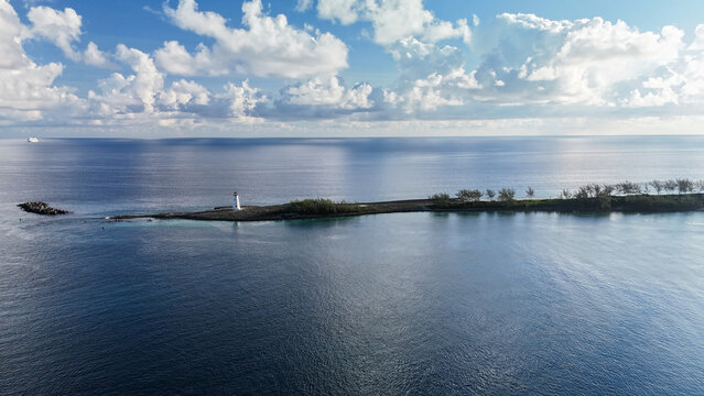 Aerial view of the lighthouse on a narrow island in Nassau Harbour, surrounded by the deep blue Caribbean Sea under a bright sky with scattered clouds in Nassau, New Providence, The Bahamas.