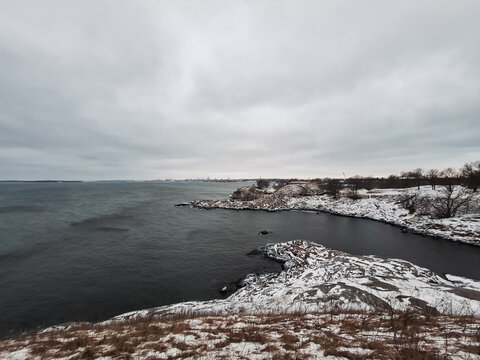 Helsinki, Finland - 02 January 2026: View of Suomenlinna fortress with snow-covered rocky coastline, dark sea waters, and a cloudy winter sky.