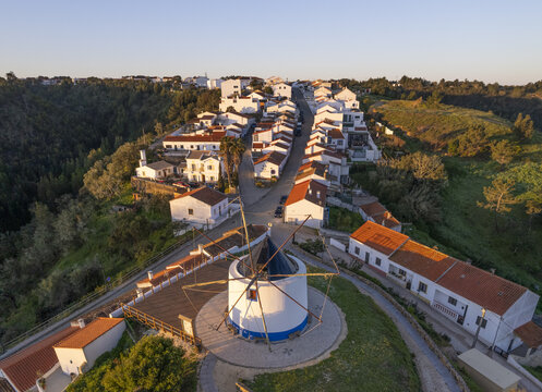 Aerial view of the historic windmill in Odeceixe, featuring white houses with terracotta roofs, winding streets, and green hills during golden hour in Odeceixe, Faro District, Portugal.