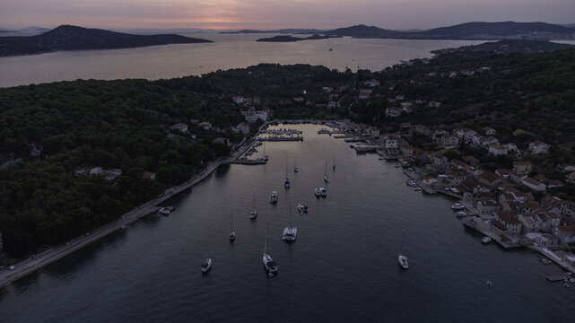 Aerial view of Kornati islands featuring boats moored in a calm bay surrounded by stone houses and dense pine forests under a soft sunset sky in Kornati, Sibensko-kninska zupanija, Croatia.