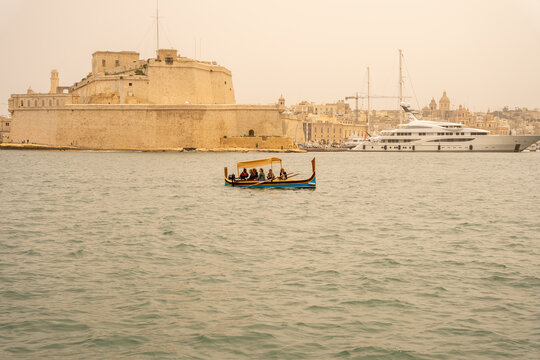 Valletta, Malta - 15 July 2022: View of a traditional dghajsa boat carrying passengers across the Grand Harbour with Fort Saint Angelo in the background.