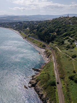 Aerial view of the Dalkey coastline featuring a train traveling along the tracks on the cliffs with lush green hills and the sea in Dublin, County Dublin, Ireland.