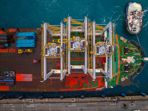 Aerial view of a cargo ship docked at the Caucedo Logistics Center with a tugboat alongside, featuring large yellow gantry structures on deck in Andres, Santo Domingo, Dominican Republic.
