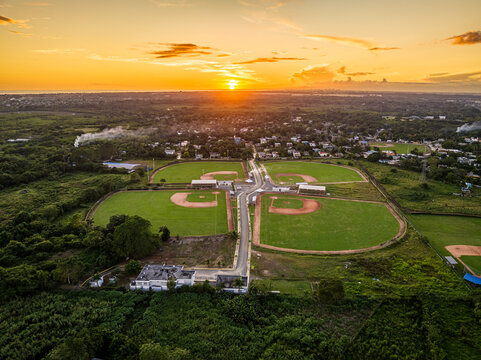 Aerial view of Quality Baseball Academy with multiple baseball fields, green grass, and dirt diamonds under a golden hour sunset in San Antonio de Guerra, Santo Domingo Provinc, Dominican Republic.