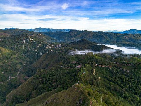 Aerial view of Little Adam's Peak surrounded by lush green mountains, tea plantations, and low-hanging clouds under a bright blue sky in Badulla, Uva Province, Sri Lanka.