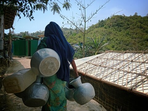 Cox's Bazar, Bangladesh - 19 July 2026: View of a woman carrying metal water pots through a refugee camp with bamboo shelters and lush green hills in the background.