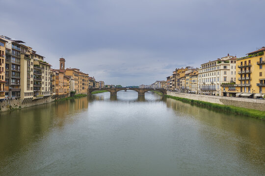 Florence, Italy - 05 May 2022: View of the historic Ponte Vecchio bridge spanning the Arno River, surrounded by traditional buildings under a cloudy sky in Florence, Italy.