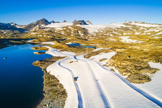Aerial view of Sognefjellet featuring a glacial lake, snow-covered mountain terrain, and tracks in the snow under a clear blue sky in Lom Municipality, Innlandet, Norway.