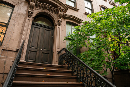 Elegant New York brownstone entrance featuring detailed stone arch, dark wooden door, and decorative iron railing. A leafy tree adds greenery to the classic urban residential facade.