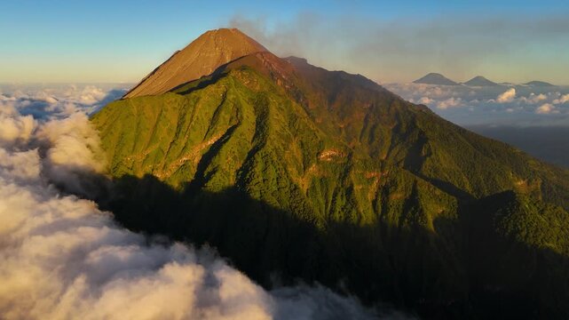 Aerial view of the lush green slopes of Merapi mountain at golden hour with clouds surrounding the volcanic peak in Cepogo, Central Java, Indonesia.
