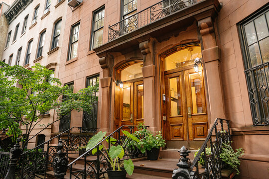 Vertical view of a historic New York brownstone facade featuring balcony details and warmly illuminated wooden doors. Decorative stonework and iron railings highlight classic urban residential