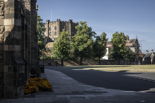 View of Durham Castle rising above the trees and stone walls, with a paved walkway in the foreground and clear blue sky in Durham, England, United Kingdom.